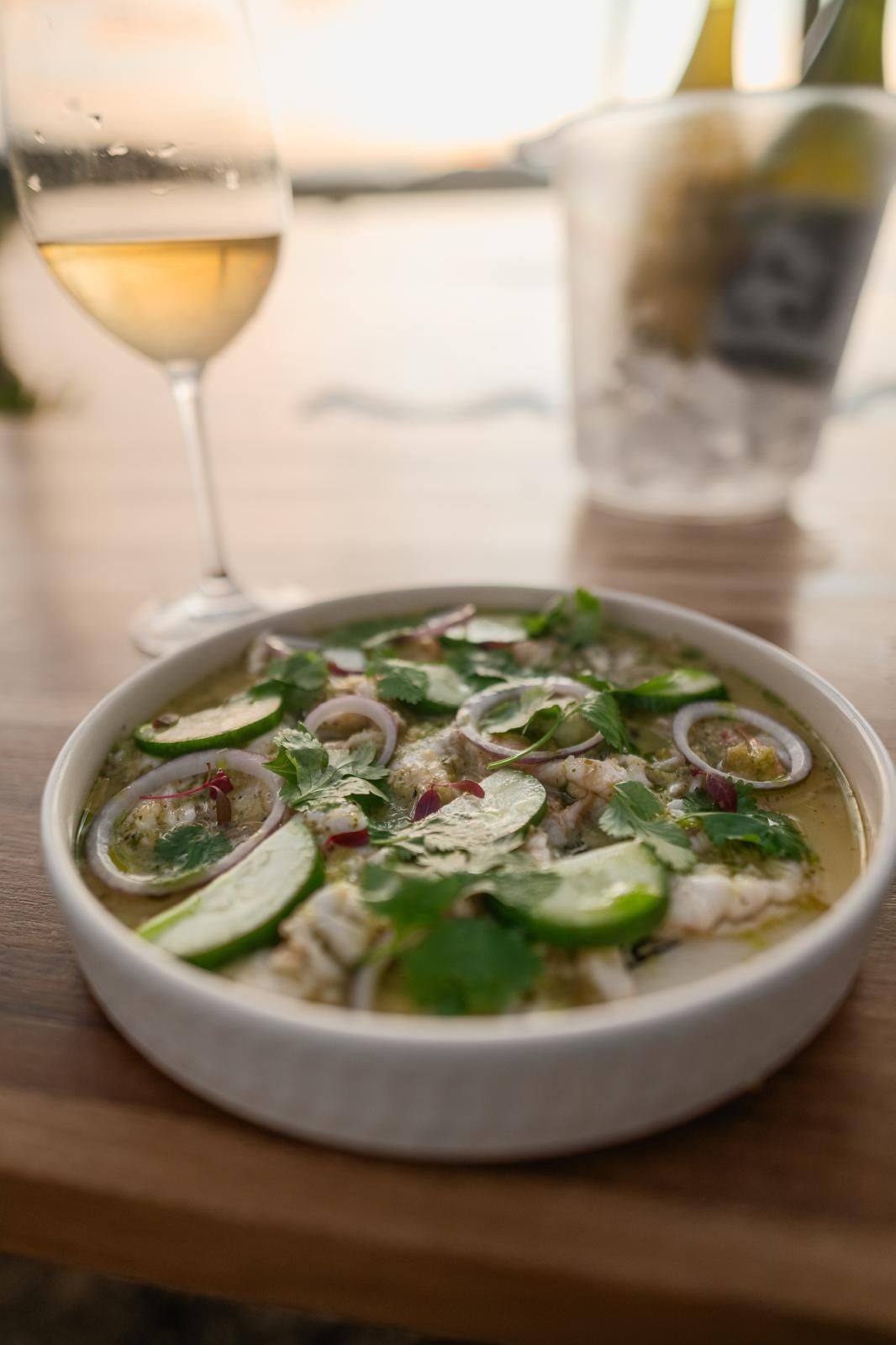 Aguachile con pepino, cebolla morada y culantro junto a una copa de vino blanco al atardecer